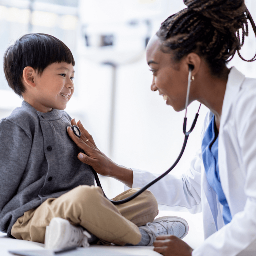 A female doctor of African decent, leans in with her stethoscope as she takes a closer listen to her patients heart. The young boy is sitting up still on the exam table.